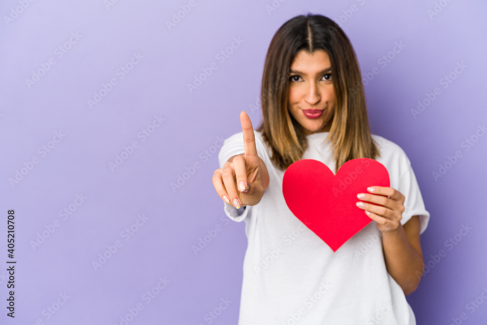 Young indian woman holding a valentines day heart isolated showing number one with finger. Young indian woman holding a valentines day heart isolated showing number one with finger.