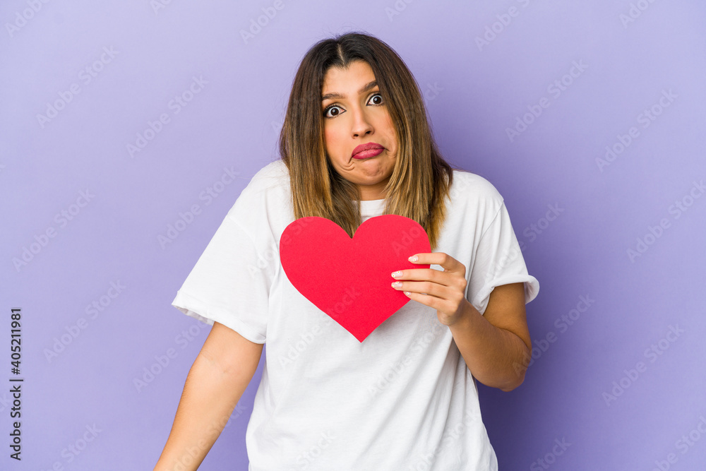 Young indian woman holding a valentines day heart isolated shrugs shoulders and open eyes confused. Young indian woman holding a valentines day heart isolated shrugs shoulders and open eyes confused.