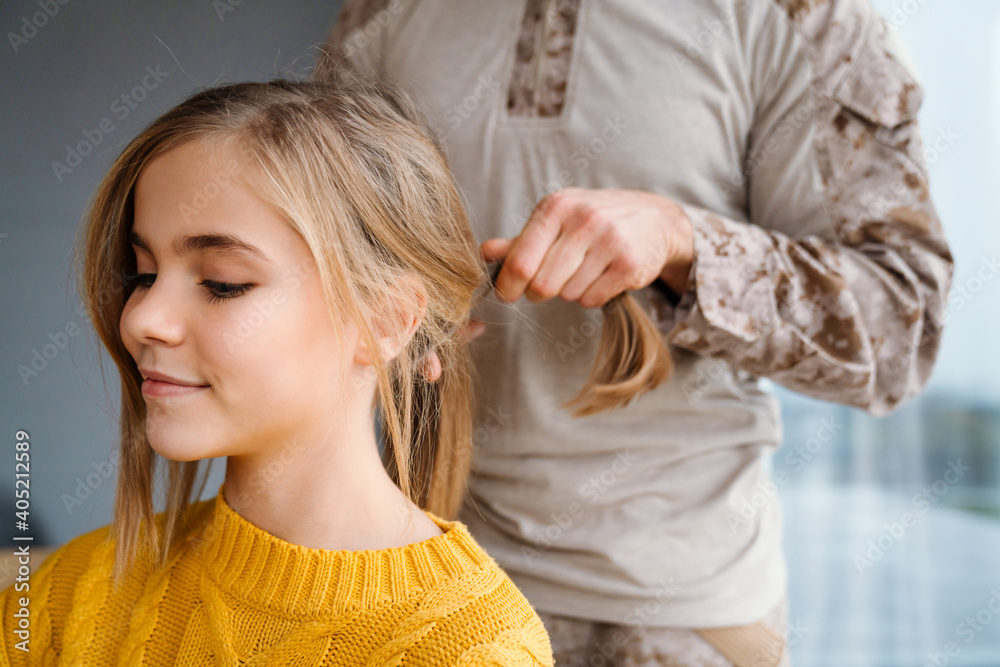 Fototapeta premium Masculine military man doing hair of his smiling daughter at home
