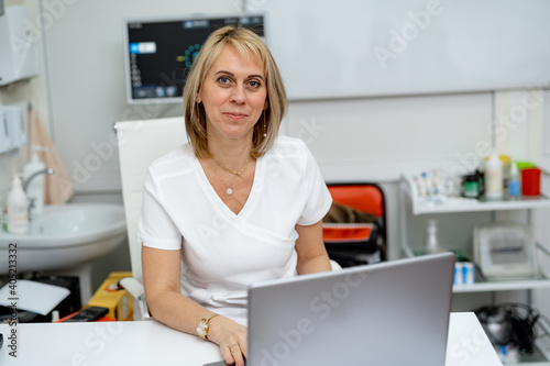 Woman doctor poses to the camera at table with laptop. Optician in scrubs. Ophthalmology concept.