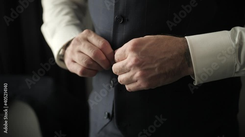 The man at the window dresses in a stylish suit and tidies himself up. The groom fastens a button on his vest. Preparing for the event. The future groom dresses up before meeting the bride.