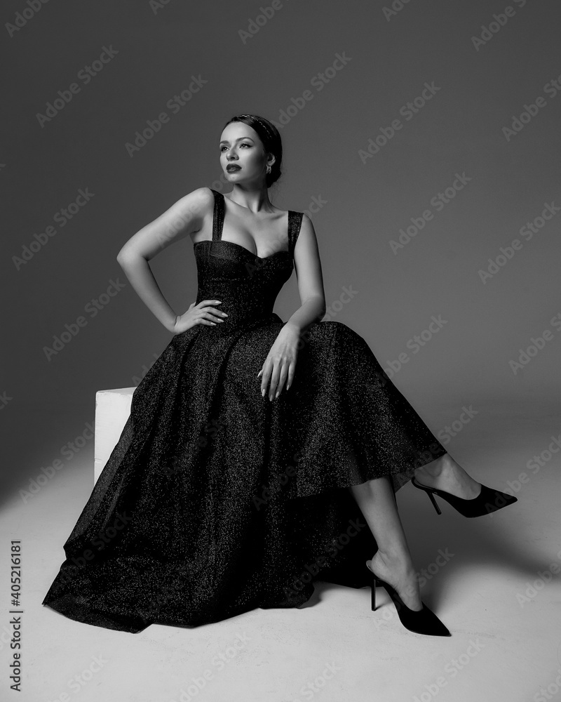 Elegant lady in black shiny ball gown and shoes sitting at white cibe with crossed legs and posing against grey background, Full length fashion portrait. Sexy female model Elegant lady in black shiny ball gown and shoes sitting at white cibe with crossed legs and posing against grey background, Full length fashion portrait. Sexy female model