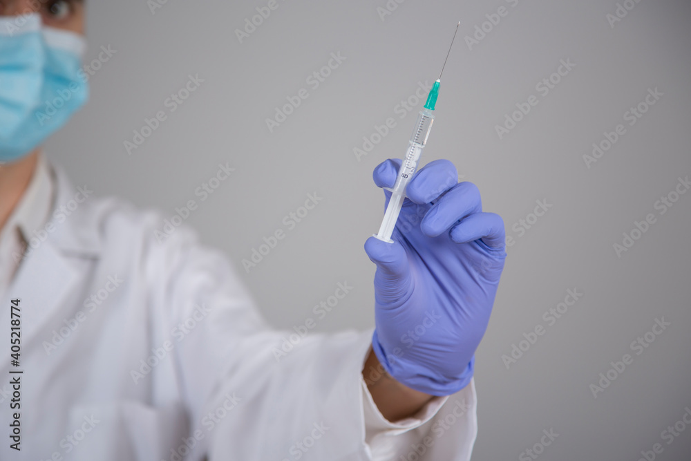 Defocused doctor in white coat and blue mask holding coronavirus vaccine syringe in hand on gray background Defocused doctor in white coat and blue mask holding coronavirus vaccine syringe in hand on gray background