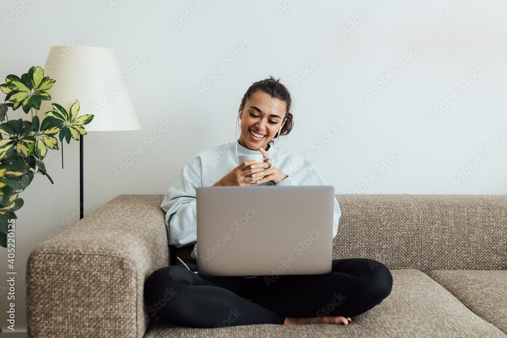 Young smiling woman holding a cup while sitting on a sofa with laptop on legs