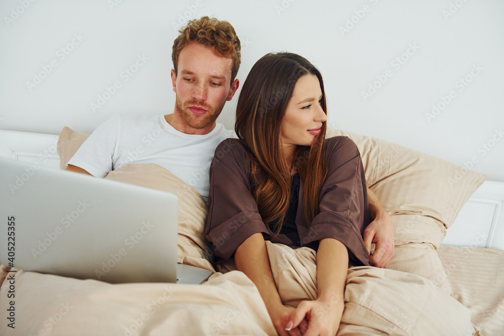 Couple lying down on bed with laptop. Interior and design of beautiful modern bedroom at daytime Couple lying down on bed with laptop. Interior and design of beautiful modern bedroom at daytime
