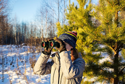 Young woman birdwatcher in winter clothes and knitted scarf looking through binoculars in winter snowy pine forest. Ecology and ornithological research, birdwatching photo