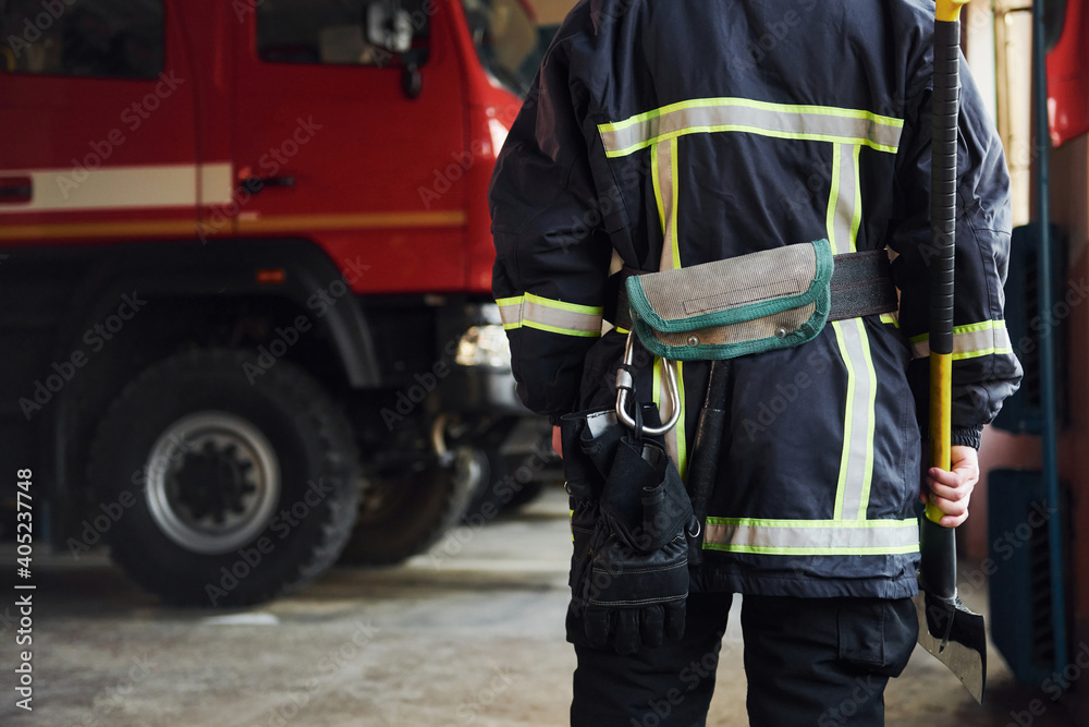 Holds axe in hands. Male firefighter in protective uniform standing near truck Holds axe in hands. Male firefighter in protective uniform standing near truck