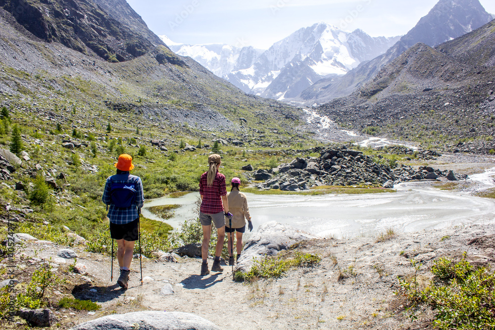 Three female hikers walk to glacier moraine by path along mountain river