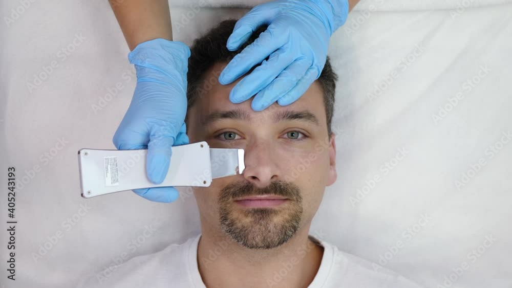 Bearded handsome middle aged man receives cosmetological ultrasonic facial cleaning, looking at camera and isolated on white background, top view. Beautician doing ultrasound peeling on male face Bearded handsome middle aged man receives cosmetological ultrasonic facial cleaning, looking at camera and isolated on white background, top view. Beautician doing ultrasound peeling on male face