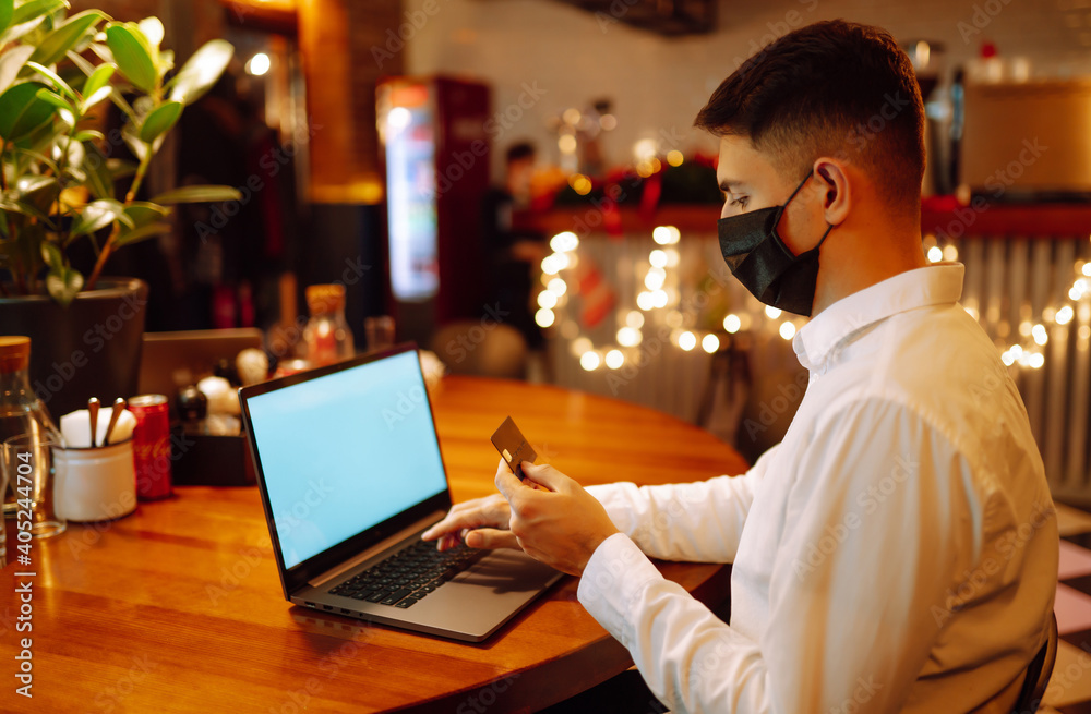 Young man in protective face mask using laptop and credit card for shopping online. Paying in internet during coronavirus quarantine. Digital payment. E-commerce store. Young man in protective face mask using laptop and credit card for shopping online. Paying in internet during coronavirus quarantine. Digital payment. E-commerce store.