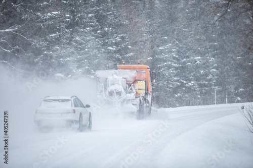 Left-handed driving road maintenance truck clears snow from the road in the forest with a car driving behind it