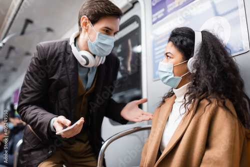 man in headphones holding smartphone and gesturing near african american woman in medical mask in subway