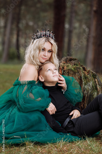 A beautiful blonde young woman in a long green dress and a diadem on her head with stylish young son in the forest. girl and boy sitting near the old stump. Solar glare. Fantasy. fairy tale