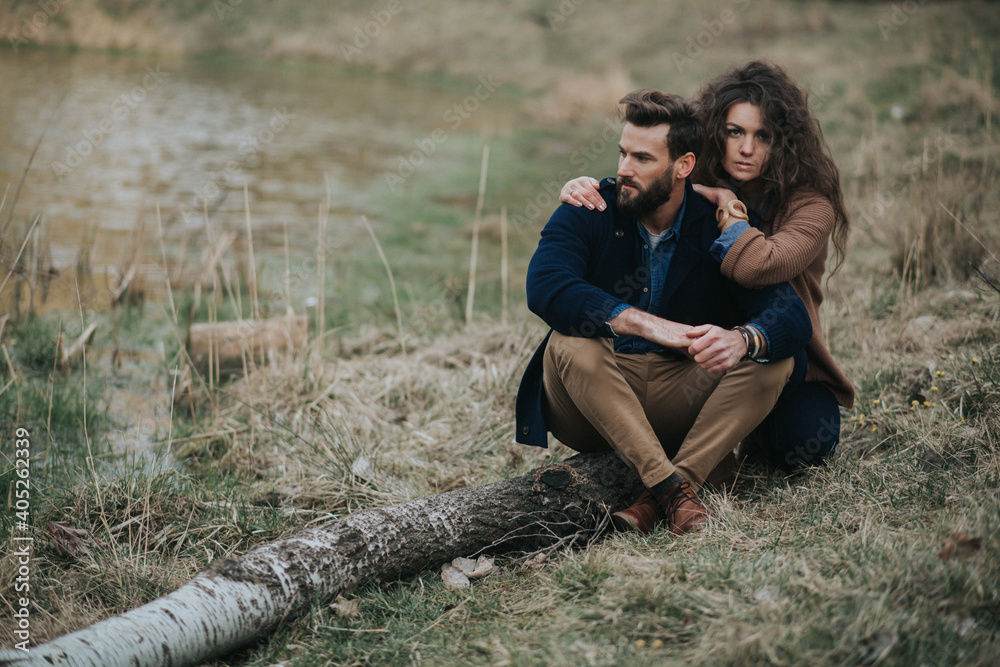 Happy caucasian lovers are sitting on the shore of the lake. Young couple is hugging on autumn day outdoors. A bearded man and curly woman in love. Valentine's Day. Concept of love and family.