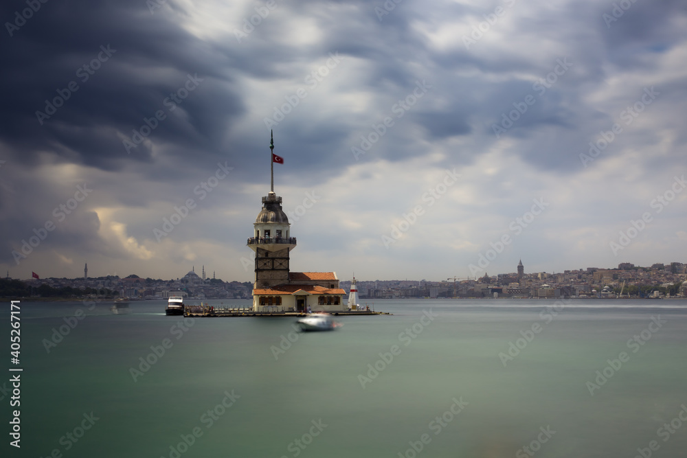 A long exposure photo of a tower looking like a lighthouse on a small island in the sea. Dramatic stormy sky and water are blurred. Oriental city in the background with mosques and minarets.