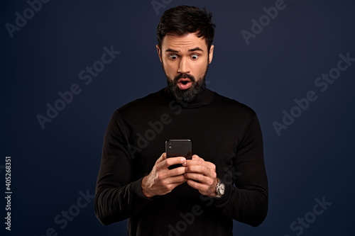 Concerned, worried young bearded man in sweater, biting lip nervously, holding smartphone indecisive, stare display, cant send risky message as feeling anxious, stand over Pacific Blue background © Roman