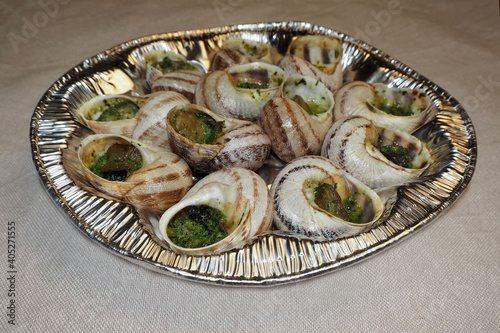 a lot of freshly prepared grape snails with green oil and garlic lies in a silver container for the oven on a beige background side view . traditional food in europe