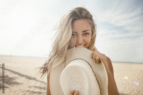Playful young woman with blond hair biting hat at beach on sunny day
