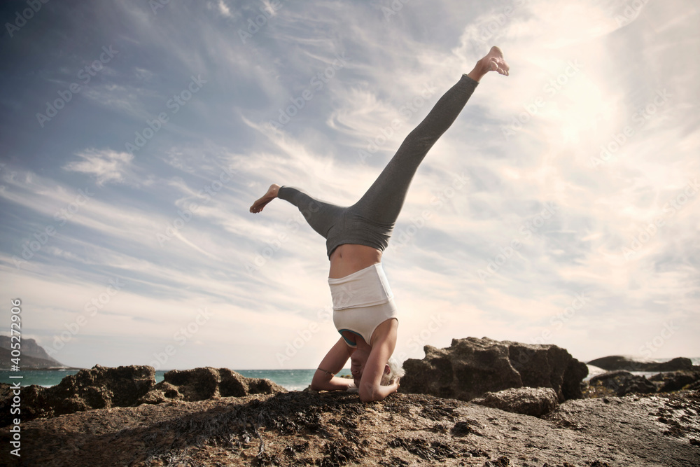 Carefree woman practicing Sirsasana with legs apart on rock formation at beach against sky Carefree woman practicing Sirsasana with legs apart on rock formation at beach against sky