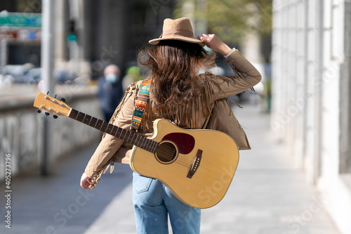 Young woman with guitar wearing hat walking on footpath in city