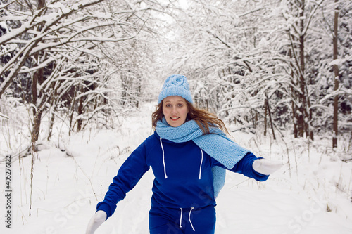 woman in a blue tracksuit white mittens and scarf stands in winter in a snow covered forest