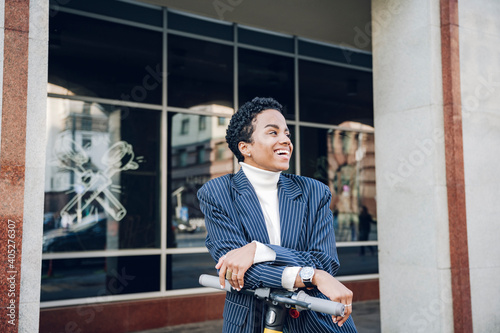 Cheerful businesswoman leaning on handlebar of electric push scooter in city