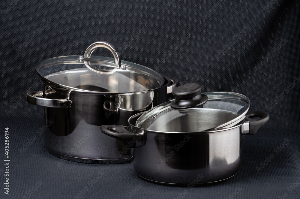Two stainless steel pans with glass lids on a dark textile background. Two stainless steel pans with glass lids on a dark textile background.