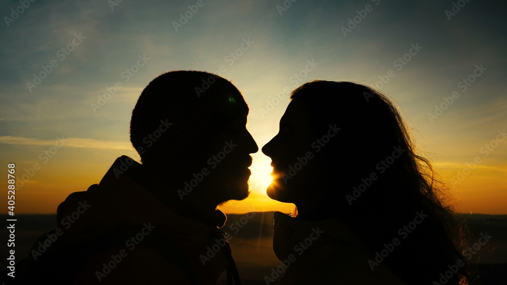 bearded man and long haired lady silhouettes kiss and laugh against back orange setting over hills sun between heads closeup