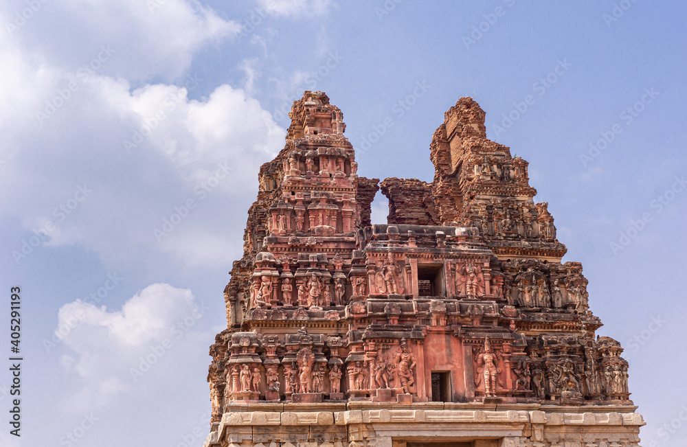 Hampi, Karnataka, India - November 5, 2013: Vittalaraya Temple. Closeup of top of ruinous red stone Gopuram with numerous statues, some sexual, under blue cloudscape.