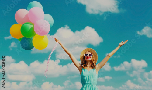 Happy smiling young woman with bunch of colorful balloons wearing a summer straw hat on the field on a blue sky background