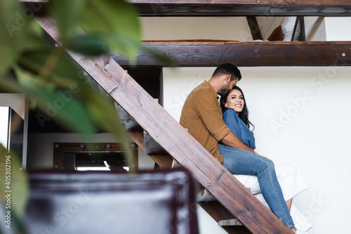 young couple embracing while sitting on wooden stairs at home, blurred foreground