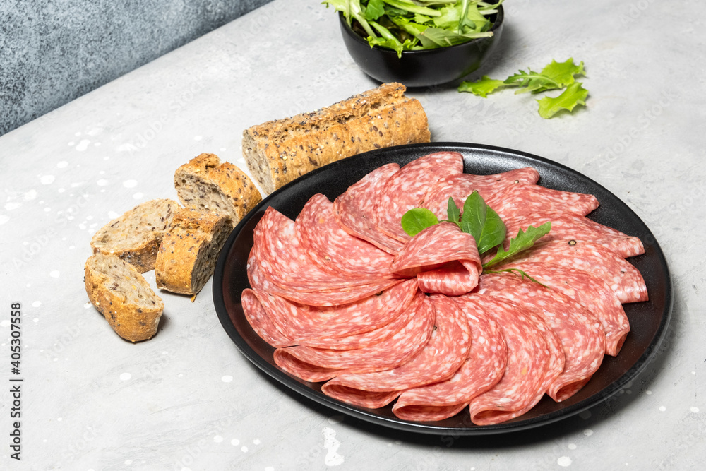 Traditional Italian salami on a black plate served with arugula and multi-grain baguette