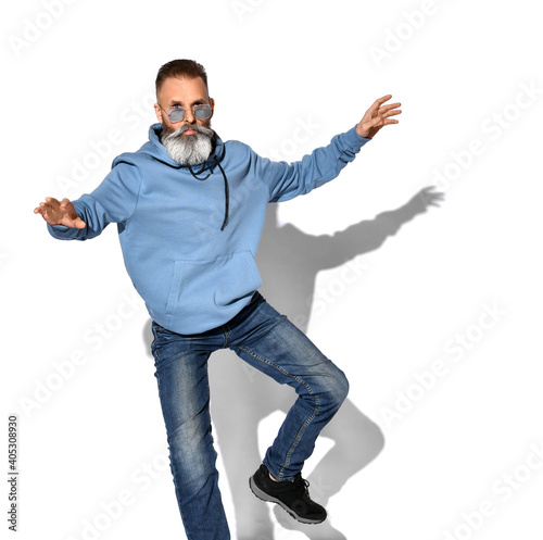 Serious gray-haired mature bearded man in trendy sunglasses, casual sweatshirt, trendy jeans, sneakers, standing or jumping with legs and arms raised. Studio shot isolated on white