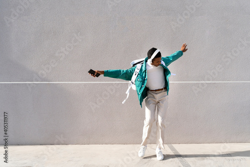 Carefree man with headphones and mobile phone dancing with arms outstretched against gray wall photo