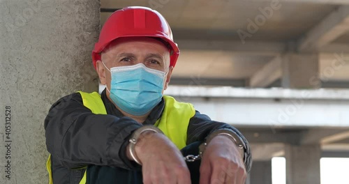 Handcuffed worker in red hard hat and protective face mask sits at construction site and looks at camera. Cinema 4K 60fps video photo