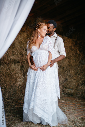 An interracial couple dressed in a rustic style is standing in a barn. African man hugs his pregnant Caucasian wife. Full-length portrait.