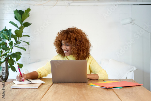 Happy businesswoman with laptop writing in notepad while working from home