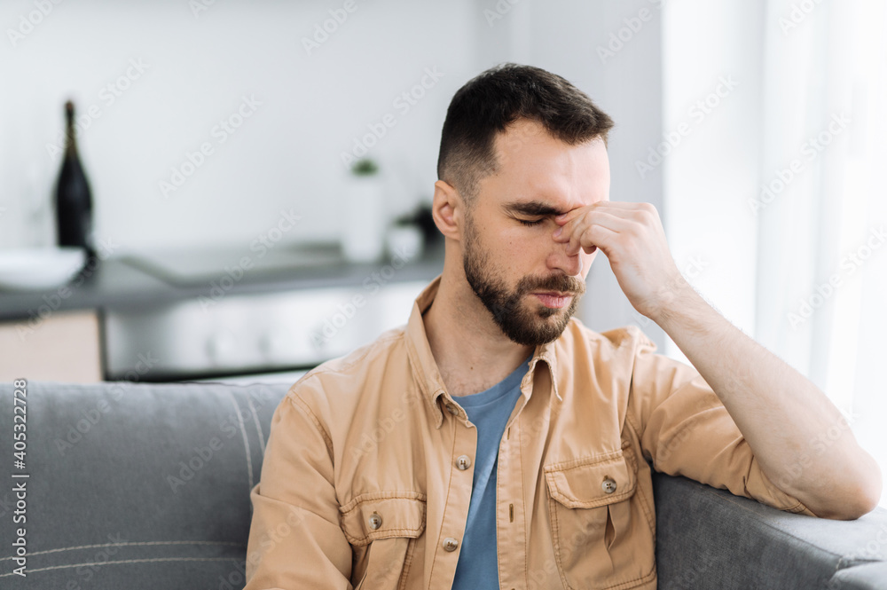 Tired caucasian bearded man sits on the sofa, having a headache. Unhealthy guy catch a cold or having flu, upset about illness, unhappy man feel depressed Tired caucasian bearded man sits on the sofa, having a headache. Unhealthy guy catch a cold or having flu, upset about illness, unhappy man feel depressed