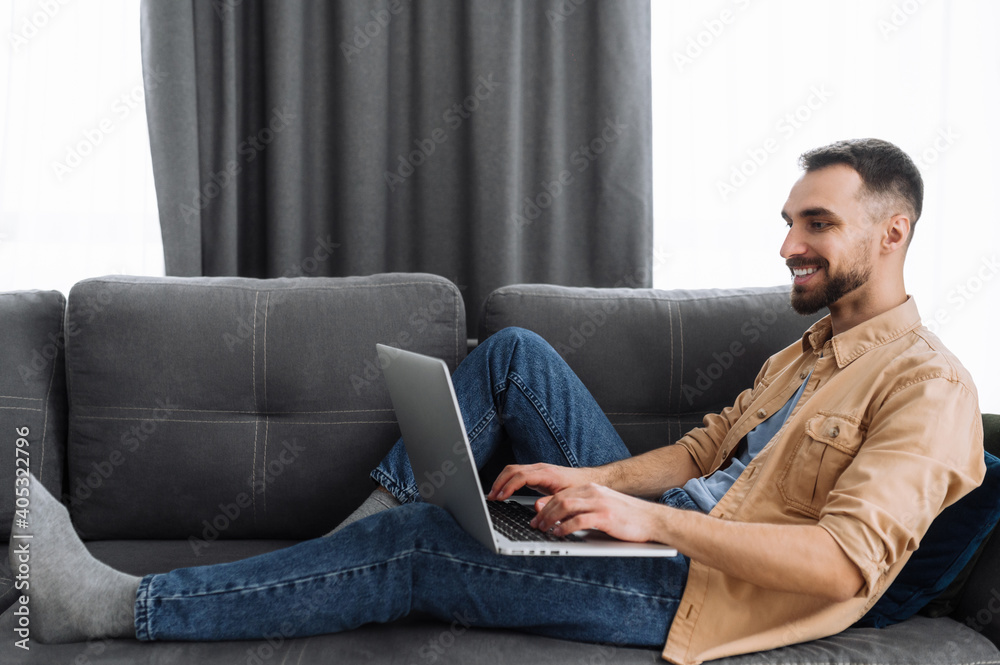 Caucasian male freelancer sits on the sofa, chilling at home. Bearded guy in casual wear use a laptop, watching films or funny videos, or working from home and smiling
