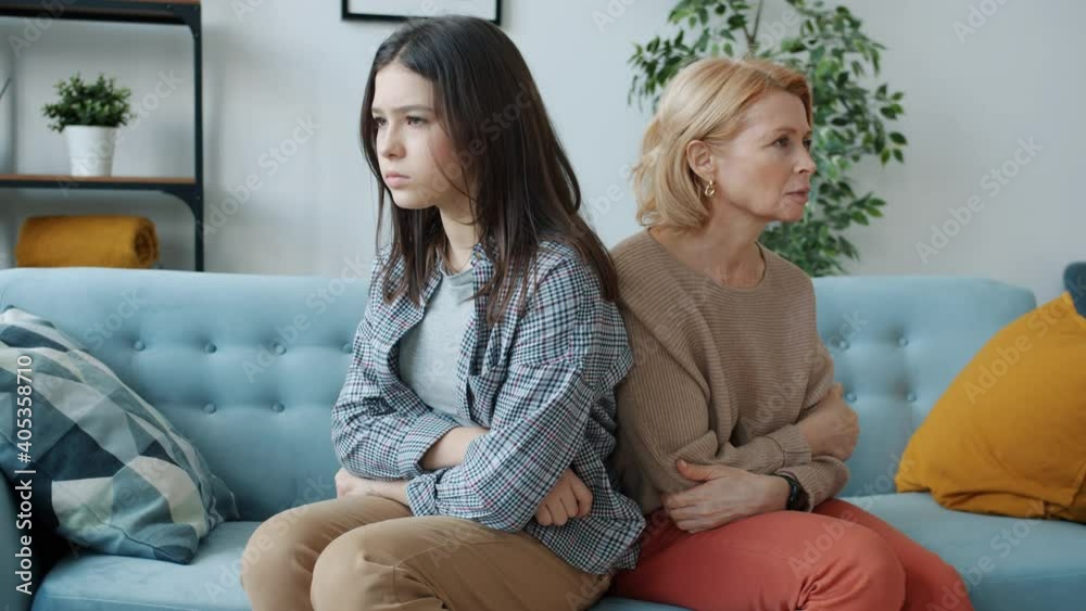 Portrait of angry mom and daughter sitting on couch back to back with arms crossed feeling mad and offended. Family relations and negative emotions concept.