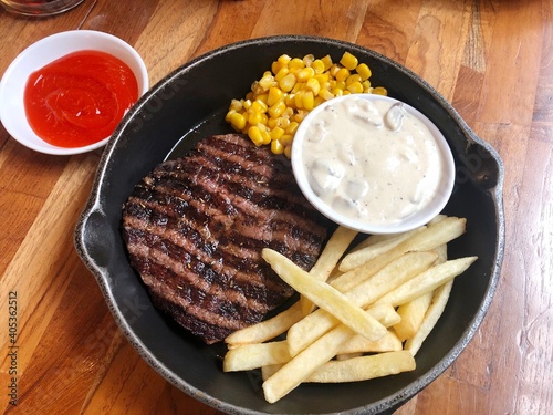Grilled sliced of well done beef steak with mushroom sauce, French fries and sauce corn on hotplate pan. Fork and spoon and chili sauce side the table. Flat lay style