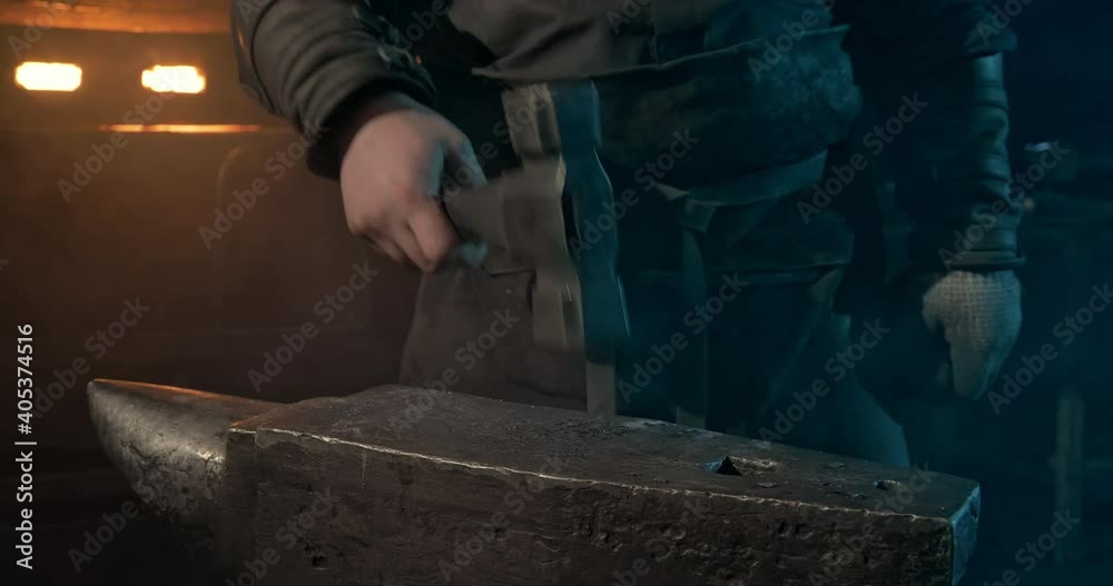 In the close-up, a large iron anvil against the background of a red-hot hearth of a forge where in slow motion a silhouette of a man in a leather apron approaches and forcefully lowers a patterned In the close-up, a large iron anvil against the background of a red-hot hearth of a forge where in slow motion a silhouette of a man in a leather apron approaches and forcefully lowers a patterned