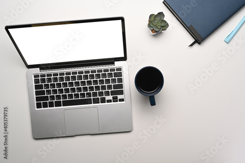 Overhead shot of contemporary workspace with mock up laptop computer, coffee cup, plant and notebook on white table.