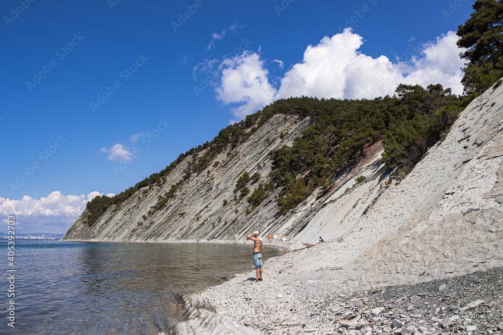 Walk along the stone wild beach at the foot of the cliffs. Beautiful summer landscape, bright blue sky with clouds, steep cliffs and a guy standing on the beach next to the campsite. Gelendzhik