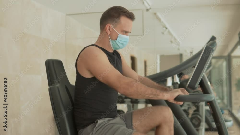 Strong male with face mask uses an exercise machine and drinks water in the gym