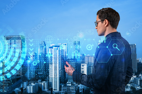 Eastern successful businessman trying to find new ideas using smartphone as a modern tool to solve problems at research and development department. Fintech hologram icons over Bangkok.