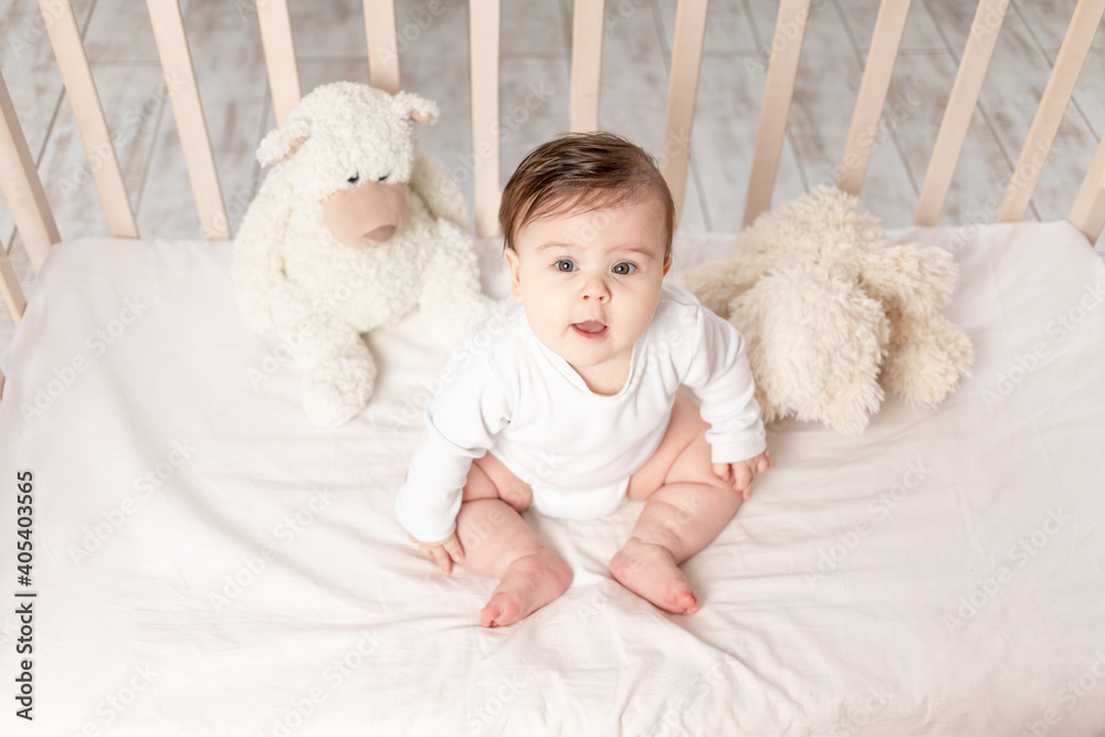 baby six months sitting in a crib in a white bodysuit with toys Teddy bears