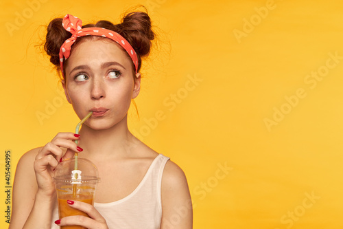 Young lady, pretty ginger woman with two buns and doted hairband. Wearing white shirt and wonder while making a sip of juicy fresh. Watching to the right at copy space over yellow background © timtimphoto