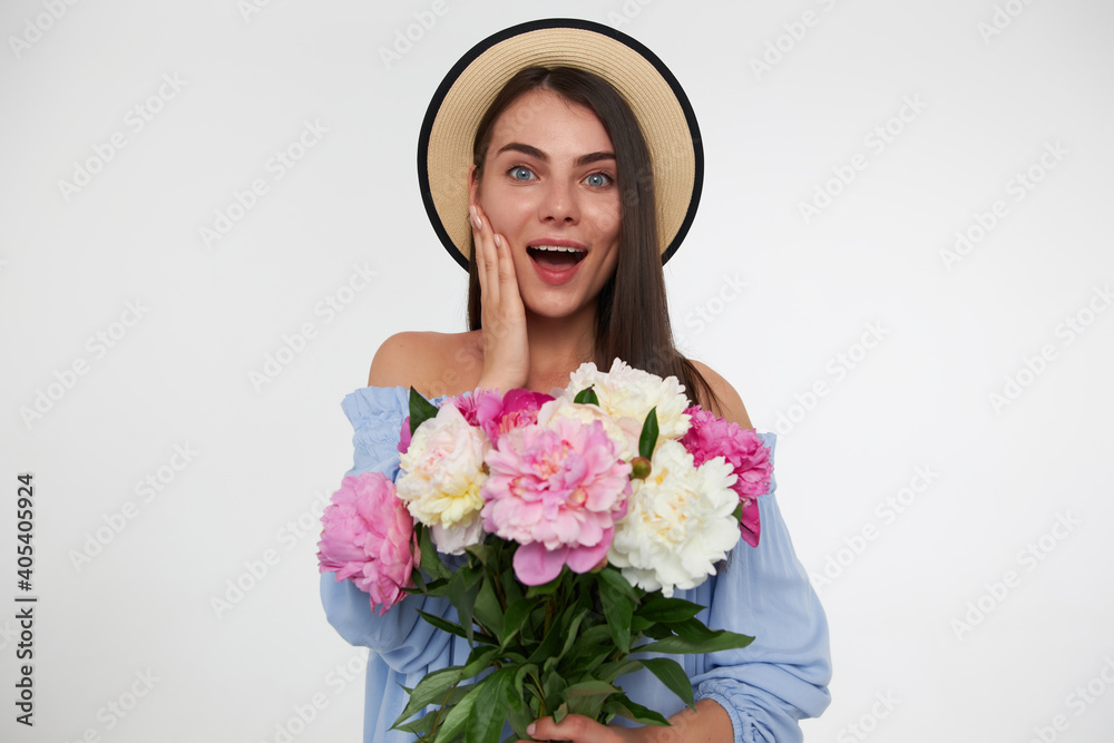 Portrait of attractive, pretty girl with long brunette hair. Wearing a hat and blue dress. Holding bouquet of flowers and touching her cheek. Watching at the camera isolated over white background Portrait of attractive, pretty girl with long brunette hair. Wearing a hat and blue dress. Holding bouquet of flowers and touching her cheek. Watching at the camera isolated over white background