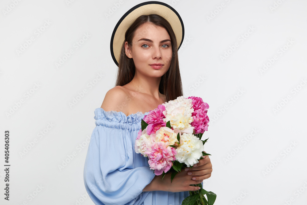Teenage girl, happy looking woman with brunette long hair. Wearing a hat and blue pretty dress. Holding a beautiful flowers. Watching at the camera isolated over white background Teenage girl, happy looking woman with brunette long hair. Wearing a hat and blue pretty dress. Holding a beautiful flowers. Watching at the camera isolated over white background